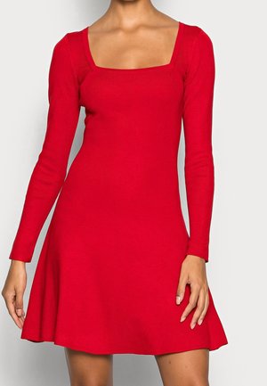 Femme portant une robe rouge à manches longues avec un col carré et une jupe évasée, debout devant un fond clair uni.