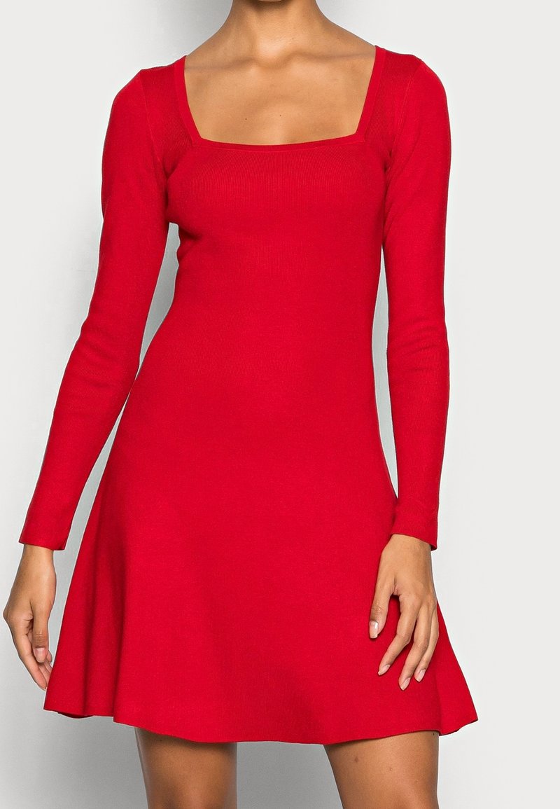 Femme portant une robe rouge à manches longues avec un col carré et une jupe évasée, debout devant un fond clair uni.