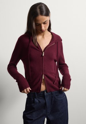 Joven mujer con una sudadera con capucha burdeos parcialmente abierta y vaqueros sueltos azul oscuro, mirando hacia abajo mientras ajusta sus pantalones con las manos.