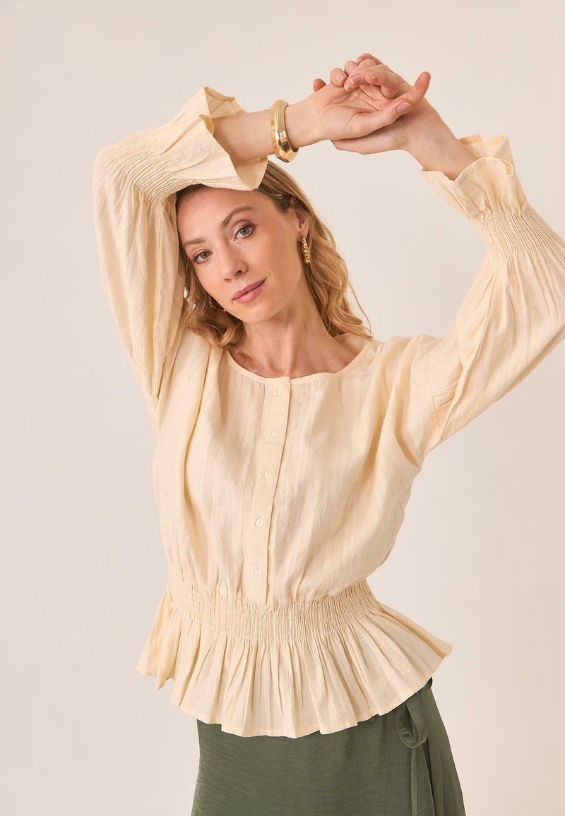 Blusa de un suave color beige claro con cuello redondo, botones en la parte delantera, mangas largas abullonadas y un dobladillo peplum fruncido. Tejido suave y texturizado.