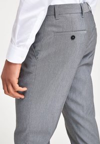 Pantalons de costume gris en tissu texturé, dotés d'une poche latérale, de passants de ceinture et d'une poche arrière avec un bouton noir, portés avec une chemise blanche.