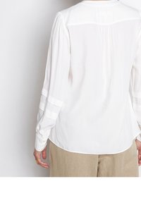 Person i en vit långärmad blus med genomskinliga horisontella ränder på ärmarna och beige byxor, sedd bakifrån.