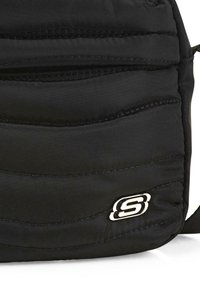 Mochila negra acolchada con textura acolchada, patrones de costuras curvadas y un accesorio de logo metálico. Incluye una correa ajustable.