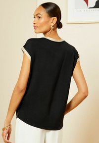 Top nero a maniche corte con scollatura rotonda e dettagli beige sulle maniche. Tessuto morbido con una vestibilità comoda, abbinato a pantaloni bianchi.