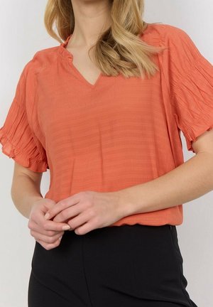 Koralowa bluzka z dekoltem w kształcie litery V z plisowanymi krótkimi rękawami, wykonana z teksturowanej tkaniny z subtelnymi pionowymi paskami, zestawiona z czarnymi spodniami.