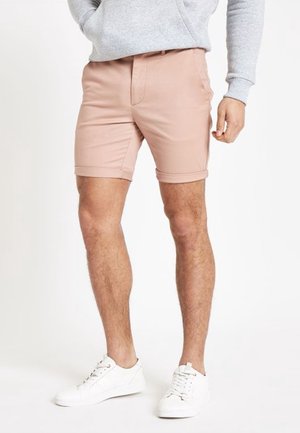 Mann, der hellrosa Shorts mit aufgerolltem Saum, grauem Sweatshirt und weißen Turnschuhen trägt, steht vor einem weißen Hintergrund.