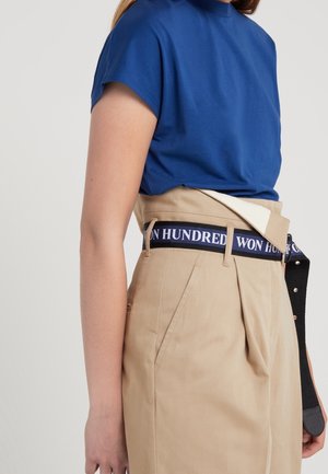 Kvinde iført en blå top med korte ærmer, stoppet ned i beige bukser med høj talje og et sort bælte med teksten "WON HUNDRED".