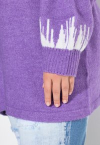 Lila Pullover mit weißem Muster am Ärmelbund, aus weichem Stoff. Gerippter Saum und Ärmelakzente. Hand liegt auf hellblauer, distressed Jeans.