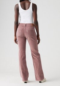 Pantalons en velours côtelé rose évasés avec taille haute, dotés de poches arrière et d'une étiquette en cuir. Portés avec un débardeur blanc.