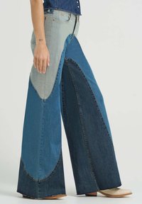 Frau trägt Jeans mit weitem Bein und Patchwork in verschiedenen Denim-Farbtönen, kombiniert mit cremefarbenen Stiefeletten, stehend vor einfarbigem Hintergrund.