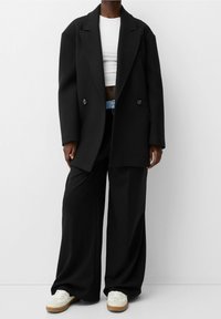 Svart dubbelknäppt blazer, vit kort topp, svarta vida byxor och vita sneakers med gummisulor, som har ett blått accentbälte.