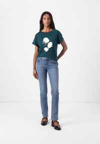 Mørkegrønn kortermet t-skjorte med hvit skjellgrafikk, kombinert med lyseblå rette jeans og svarte lakksko.