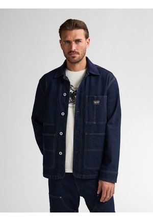 Denim jacket in dark blue with a button closure, multiple pockets, and contrasting stitching. Worn over a light-colored graphic t-shirt.