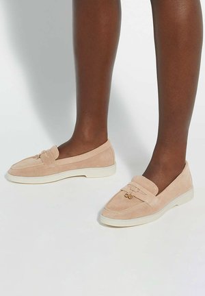 Suede loafers in light beige with a white rubber sole. Features a decorative front strap and gold-tone hardware accents.