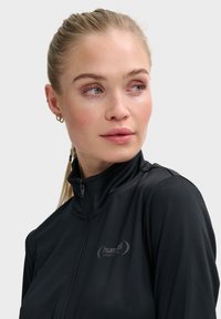 Zwarte zip-up sportjack met een hoge kraag, gemaakt van glad, rekbaar materiaal met een subtiel logo op de borst.