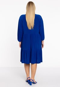 Blaues, knielanges Kleid mit langen, puffärmeln, gerafftem Taillenbereich und gestuftem Rock. Getragen mit passenden blauen Blockabsatzsandalen.