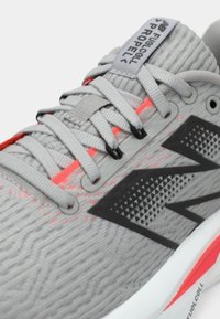 Chaussure de sport gris clair avec une tige en tissu texturé, accents orange néon et un logo noir bien visible sur le côté.