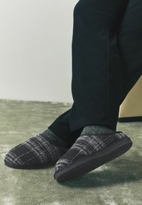 Mustat ja harmaat ruudulliset tossut, joissa on pehmeä päällyskangas ja teksturoitu kumipohja, käytössä tummien housujen ja harmaiden sukkien kanssa vaalealla pinnalla.
