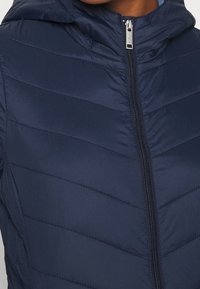 Granatowa ocieplana kurtka z kapturem, wyposażona w zamek błyskawiczny z przodu, z przeszyciem w kształcie jodełki oraz gładką, błyszczącą fakturą.