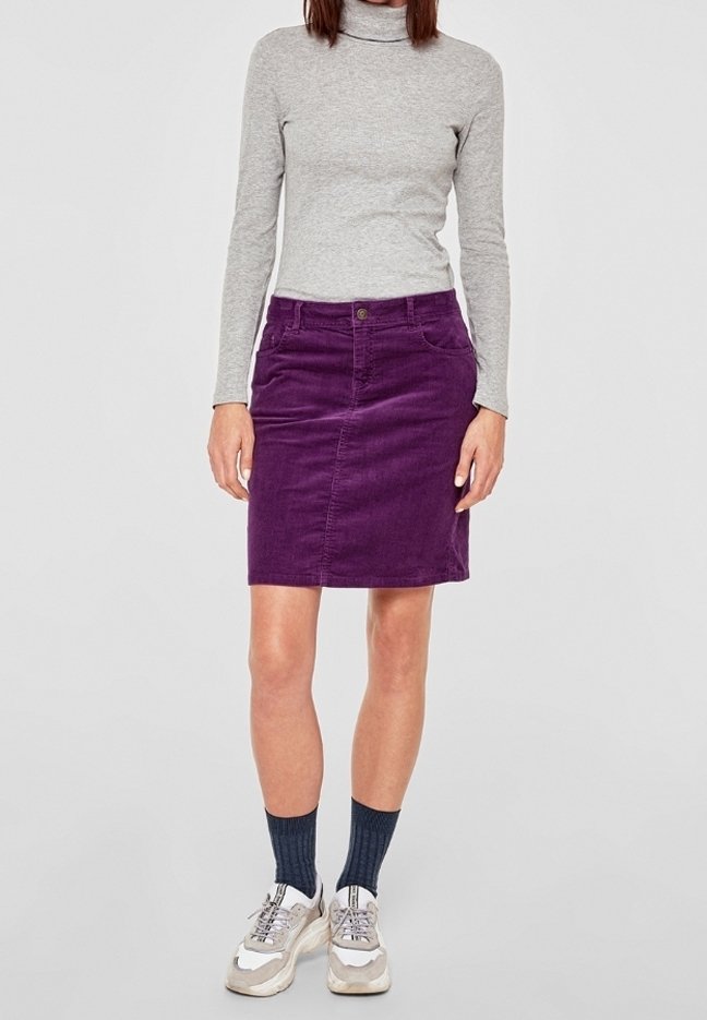 Personne portant un col roulé gris à manches longues, une jupe violette, des chaussettes foncées et des baskets blanches, debout devant un fond uni.