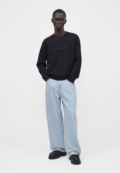 Homme debout, les mains dans les poches, portant un sweat-shirt noir Emporio Armani, un jean large bleu clair et des chaussures noires.