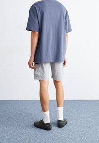 Hombre de pie sobre una alfombra azul, vistiendo pantalones cortos grises, una camiseta azul de gran tamaño, calcetines blancos y zapatillas de deporte de color verde oscuro, de espaldas a la cámara.