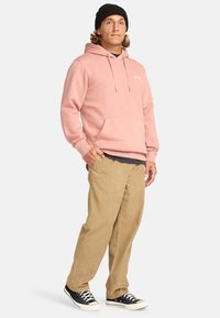 Light pink hoodie with a kangaroo pocket, black beanie, beige corduroy pants, and black sneakers with white soles and laces.