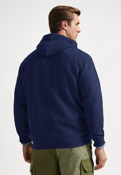 Vyras, dėvintis tamsiai mėlynę bliuzoną su gobtuvu ir žalias kelnes su kišenėmis, pavaizduotas iš nugaros, stovintis prieš baltą foną.