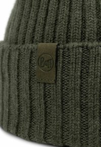 Dark green knitted beanie with a ribbed texture and a fabric label featuring a circular logo. Material appears soft and thick.