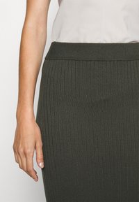 Dark green ribbed knit skirt with a elastic waistband, close-fitting and smooth texture, paired with a light top, showcasing a hand.