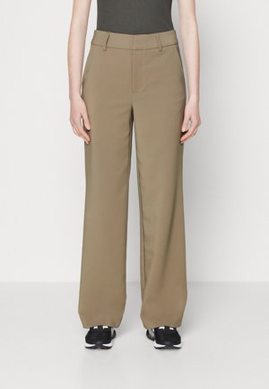 Person wearing high-waist, wide-leg beige trousers with belt loops and black sneakers, standing against a plain white background.