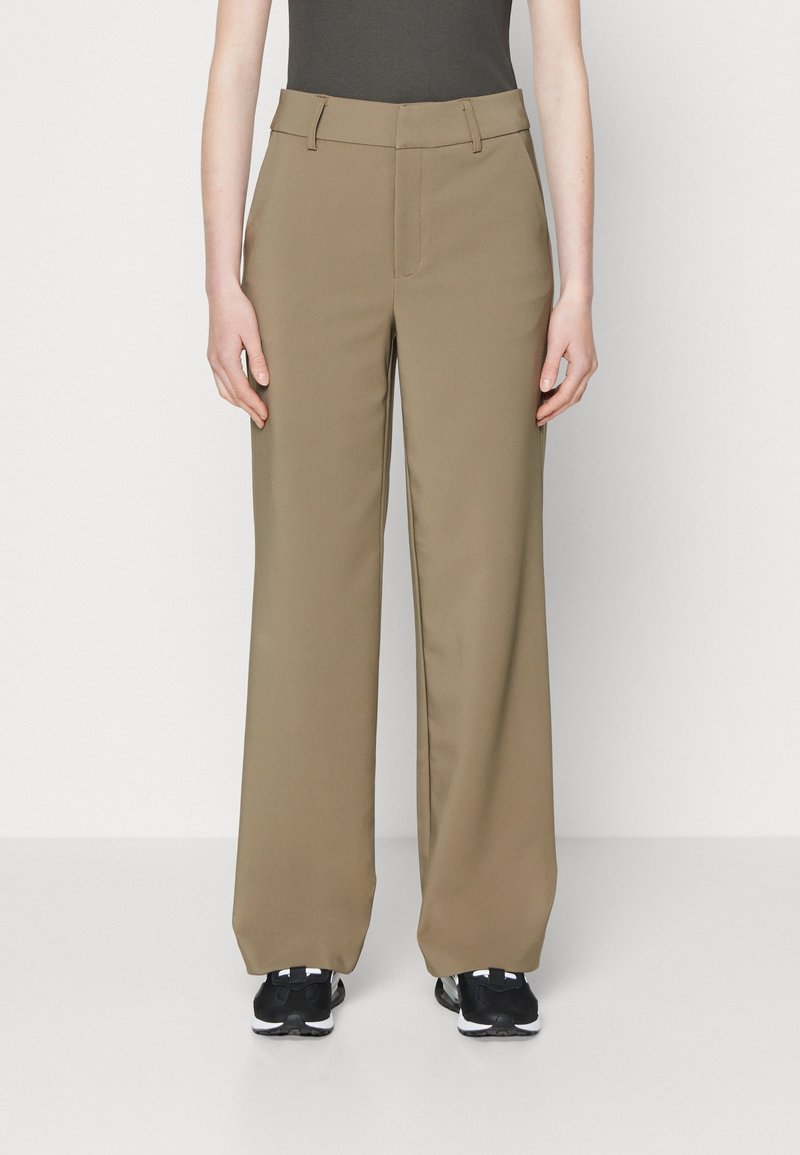 Personne portant un pantalon beige taille haute à jambes larges avec passants de ceinture et baskets noires, debout devant un fond blanc uni.