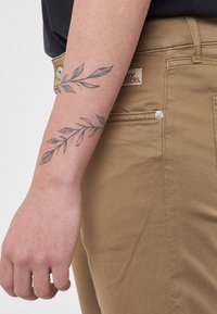 Pantaloni marroni chiari realizzati in tessuto testurizzato, con una tasca laterale e un'etichetta del marchio. È visibile un braccio con un tatuaggio di rami di foglie.