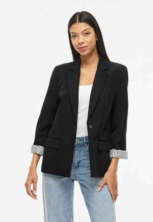 Vrouw die een zwarte blazer draagt met opgerolde gestreepte manchetten, een wit topje en lichtblauwe jeans, staand tegen een effen witte achtergrond.