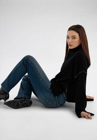 Mujer de cabello largo y castaño sentada en el suelo, vistiendo un suéter de cuello alto negro, jeans azules y botas negras con tacón, mirando hacia la cámara.