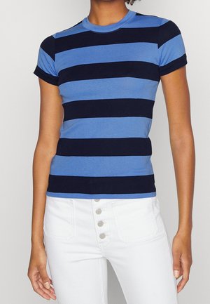 Personne portant une chemise bleu et noir à rayures horizontales et manches courtes ainsi qu’un pantalon blanc taille haute avec des boutons visibles à l’avant.