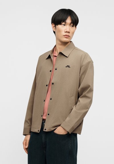 Άνδρας με μπεζ σακάκι με κουμπιά πάνω από ροζ πουκάμισο, με το χέρι στην τσέπη, κοιτάζοντας προς τα αριστερά του σε απλό φόντο.