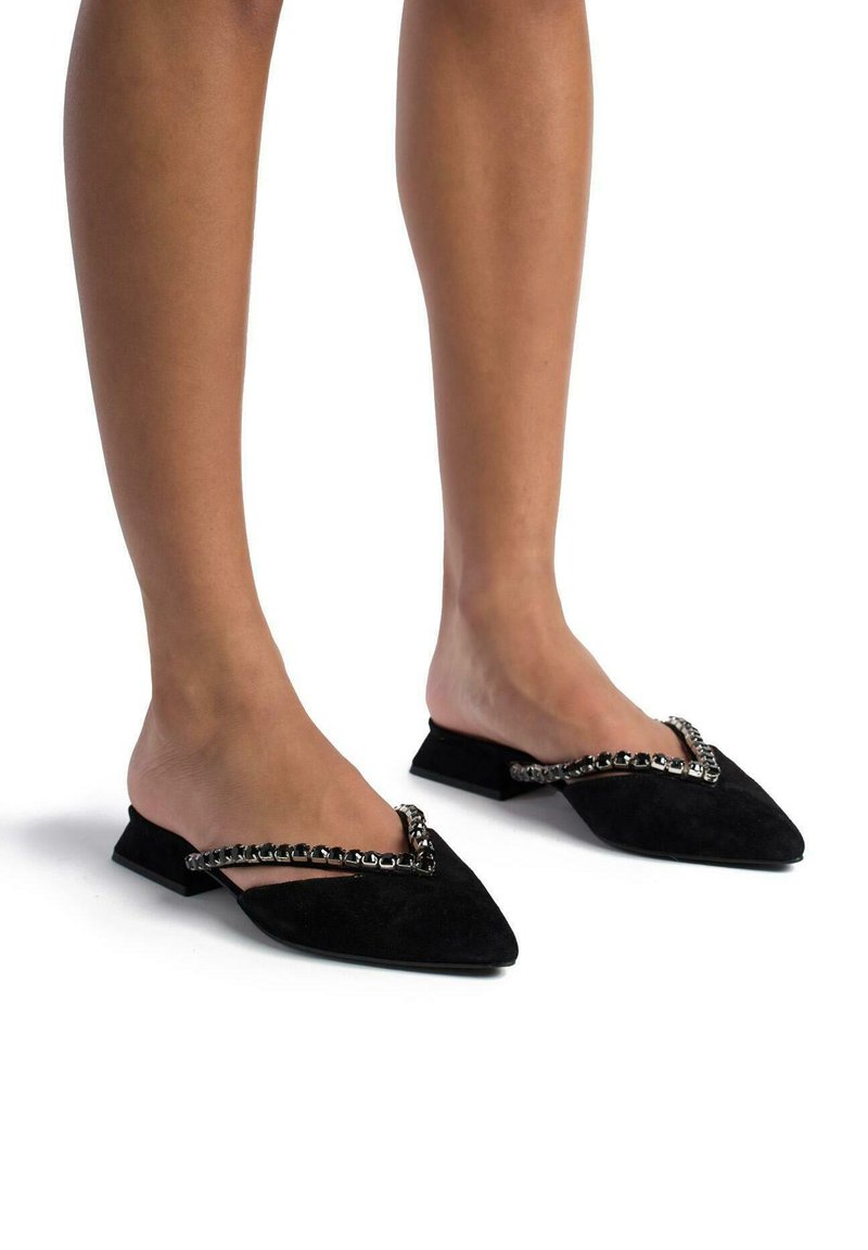 Mule in suede nero con punta affusolata e tacco piatto, caratterizzato da un accentuato da una catena metallica lungo l'apertura per un tocco di design in più.