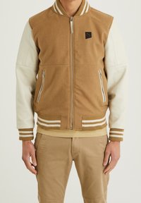 Brązowa bomberka z wełny z kremowymi skórzanymi rękawami, dwiema kieszeniami na zamek, prążkowanymi ściągaczami na mankietach oraz naszywką z logo na piersiach.