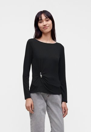 Femme souriante aux longs cheveux foncés portant un top noir à manches longues avec un nœud sur le côté et un jean gris clair sur fond blanc.