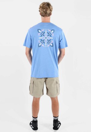 Homme portant un t-shirt bleu clair avec un motif floral rose et bleu foncé dans le dos, un short cargo beige, des chaussettes noires et des baskets noires, debout de dos.