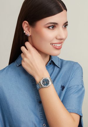 Montre en argent avec un cadran bleu et des accents en diamant, portée au poignet. Chemise en denim bleu à manches courtes, agrémentée de boucles d'oreilles en clou.