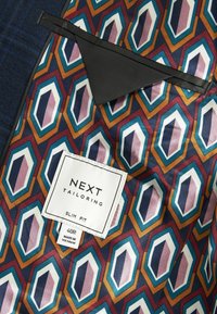 Chaqueta slim fit de Next con forro que presenta un patrón geométrico en azules, naranjas y blancos, con un bolsillo negro y etiqueta de cuidado.