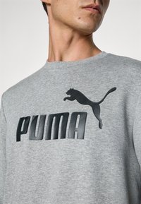 Homme portant un sweat-shirt gris clair avec un grand logo Puma noir et un emblème de chat sautant sur la poitrine, légèrement tourné vers la droite.