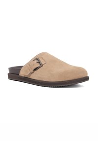 Beige suede slip-on shoe with a smooth texture, rounded toe, and a metal buckle accent on the side. Flat, dark rubber sole.