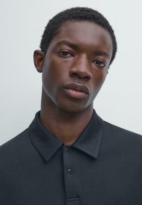 Polo noir avec col classique, patte de boutonnage et texture lisse. Le modèle a les cheveux courts et naturels, avec une expression neutre.