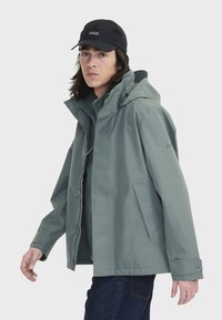 Veste verte déperlant avec capuche, boutons-pression et poches latérales. Portée par-dessus une couche intérieure plus foncée et associée à un jean noir.