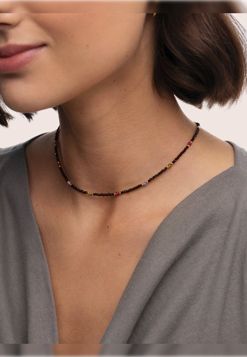 Primer plano de una mujer que lleva un delicado collar tipo choker oscuro con pequeñas cuentas multicolores y una camiseta de escote en V gris.