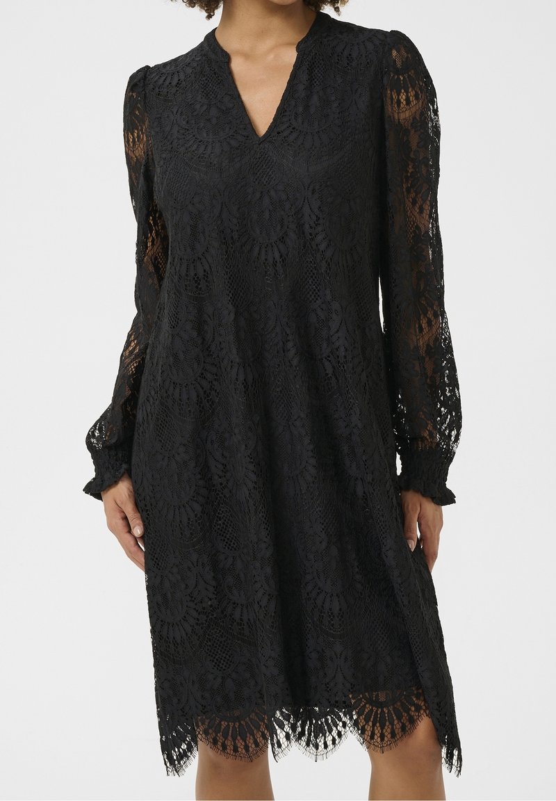 Robe noire en dentelle à manches longues, col en V, ourlet festonné et manches transparentes à motifs. Modèle présenté du cou aux genoux sur fond uni.