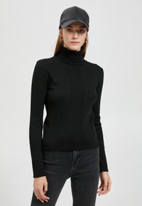 Schwarzer gerippter Rollkragenpullover mit langen Ärmeln, kombiniert mit dunklen Jeans und einer schwarzen Ledermütze, die eine figurschmeichelnde Silhouette bietet.